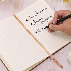 May include: Open notebook with lined pages and a brown leather cover. The text "Call Grandma", "Grocery shopping", and "Don't" are written on the page. A black pen is held over the page, ready to write. A brown ribbon bookmark is visible.