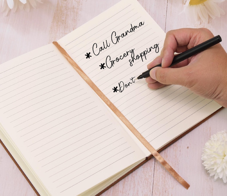 May include: Open notebook with lined pages and a brown leather cover. The text "Call Grandma", "Grocery shopping", and "Don't" are written on the page. A black pen is held over the page, ready to write. A brown ribbon bookmark is visible.