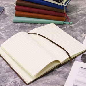 May include: An open, lined notebook with a brown cover and a brown ribbon bookmark. A stack of similar notebooks in various colors, including brown, red, and blue, is visible in the background. The pages are cream-colored.