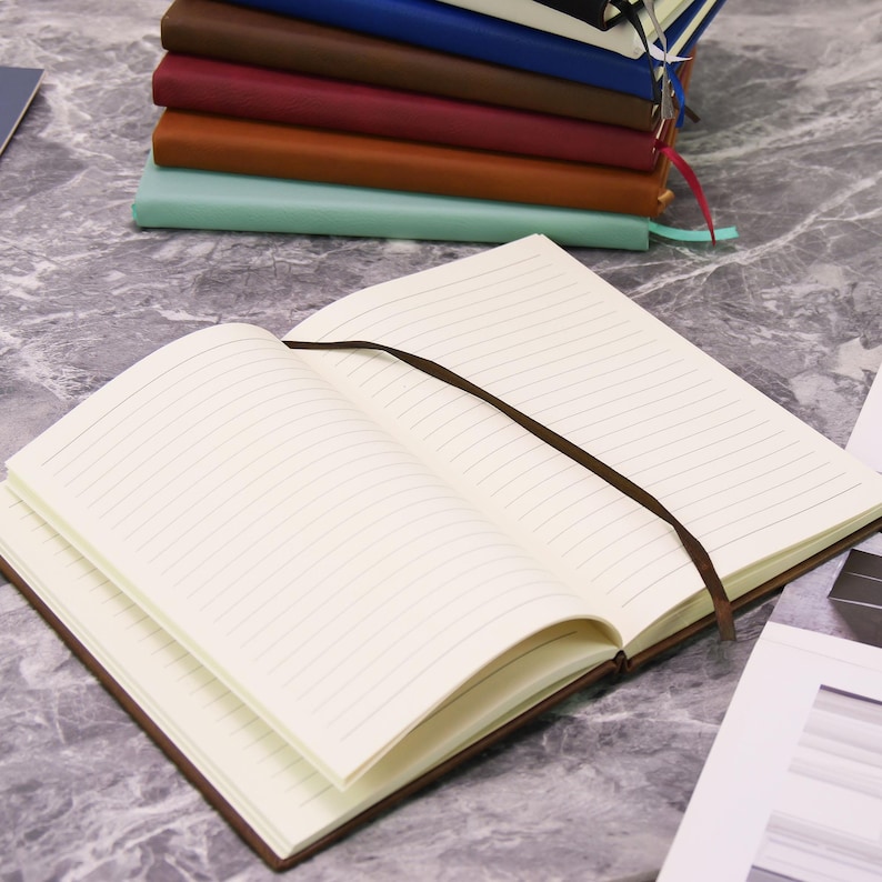 May include: An open, lined notebook with a brown cover and a brown ribbon bookmark. A stack of similar notebooks in various colors, including brown, red, and blue, is visible in the background. The pages are cream-colored.