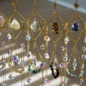 May include: A gold-colored geometric mobile with hanging crystals and gemstones. The mobile features a sunburst design at the top and diamond-shaped frames holding various crystals. Additional crystals and decorative elements dangle from chains below.