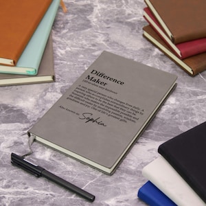 May include: A gray notebook with the text "Difference Maker" and a definition. The notebook has a ribbon bookmark and the name "Sophia" below the definition. Other notebooks and a pen are also visible.
