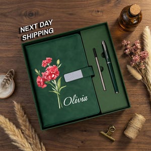 May include: A dark green notebook with a floral print of red carnations and the name Olivia printed in white. The notebook features a matching green strap with a silver nameplate reading Olivia, accompanied by a matching pen in a gift box.