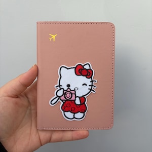 Puede incluir: Un tarjetero rosa con un personaje de dibujos animados con un lazo y vestido rojos, sosteniendo una cámara. Un icono de avión amarillo está en la esquina superior izquierda. El tarjetero es de material liso.