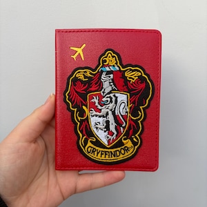 May include: Red passport holder with the Gryffindor crest and a small yellow airplane. The crest features a lion, a crown, and the word "Gryffindor". The passport holder is made of a smooth material.