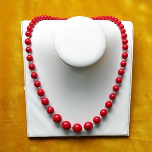 May include: A red beaded necklace on a white display stand. The necklace is made of round, red beads.