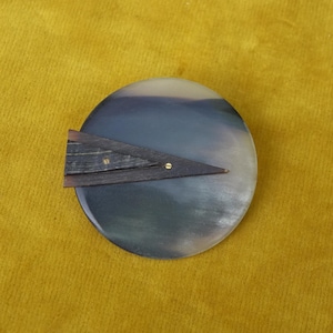 Könnte beinhalten: Ein rundes Objekt in Dunkelgrau und Creme mit einem dunkelbraunen, dreieckigen Holzakzent. Das Holz ist mit kleinen Messingnieten befestigt. Das Objekt befindet sich vor einem gelben Hintergrund.