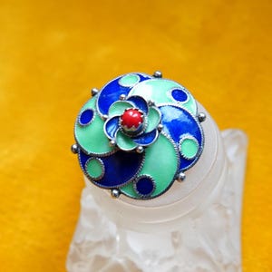 Puede incluir: Broche circular con diseño floral. El broche presenta una piedra roja central rodeada de pétalos azules y verdes, con detalles plateados. El broche se encuentra sobre un fondo amarillo.