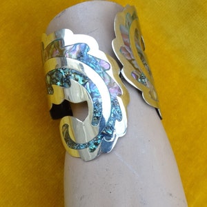 May include: A silver bracelet with a decorative design featuring iridescent abalone shell inlay. The bracelet is open and has a wide, flat band.