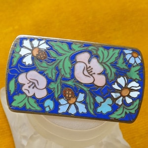 May include: A rectangular enamel brooch with a blue background and a floral design. The flowers are pink, white, and green.