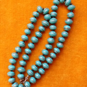 May include: A turquoise blue beaded necklace with silver accents. The beads are round and have a striped pattern. The necklace is on a bright orange background.