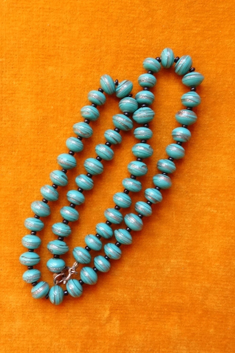 May include: A turquoise blue beaded necklace with silver accents. The beads are round and have a striped pattern. The necklace is on a bright orange background.