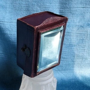 Könnte beinhalten: Eine kleine, rechteckige, burgunderfarbene Lederbox mit einem Scharnierdeckel und einem kleinen, klaren Glasspiegel auf der Vorderseite.