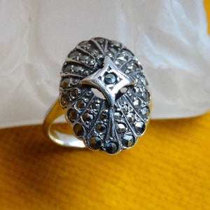 Op de afbeelding: Een zilveren ring met een gefacetteerde zwarte steen gezet in een geometrisch ontwerp. De ring is gemaakt van sterling zilver en heeft een gedetailleerd ontwerp.