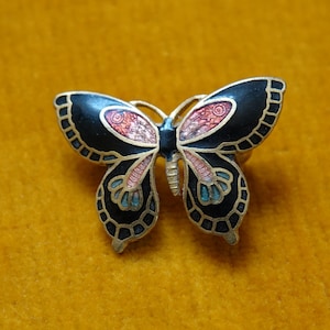 May include: A butterfly-shaped brooch featuring black enamel wings outlined in gold-tone metal. The wings are decorated with intricate patterns in shades of pink, orange, and light blue. The central body of the butterfly is also gold-toned.