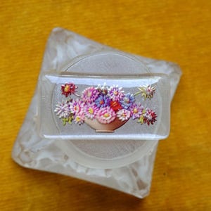 Könnte beinhalten: Transparente, rechteckige Vintage-Brosche mit floralem Design. Die Brosche zeigt einen Strauß bunter Blumen in einer Vase. Die Brosche ist auf einem weißen, marmorierten Sockel angebracht.