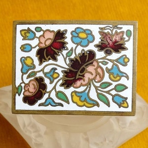 May include: A brass rectangular box with a white enamel top featuring a floral design in shades of pink, red, blue, yellow, and green.