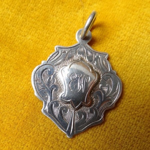 May include: Silver pendant with an ornate design and a monogrammed initial "E" in the centre.