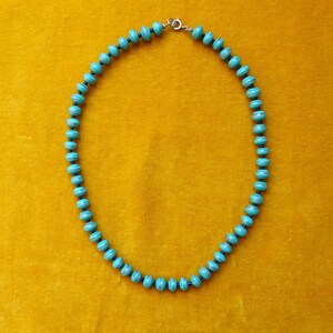 May include: A turquoise blue necklace with a silver clasp, made of round beads with black accents. The necklace is on a yellow background.
