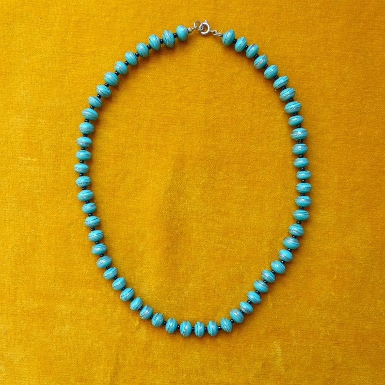 May include: A turquoise blue necklace with a silver clasp, made of round beads with black accents. The necklace is on a yellow background.
