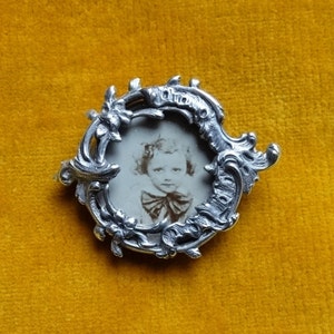 Puede incluir: Un medallón de plata con un diseño floral ornamentado. El medallón contiene una fotografía en blanco y negro de un niño pequeño.