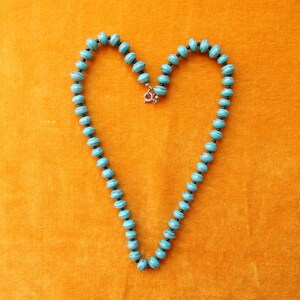 May include: A turquoise and silver necklace with round beads, shaped into a heart on a yellow background.