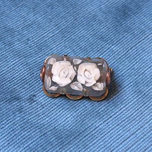 May include: A rectangular brooch with a clear, possibly glass, body. Two white rose carvings are visible inside, along with white leaf carvings. The brooch is framed by a gold-colored metal with a decorative design.