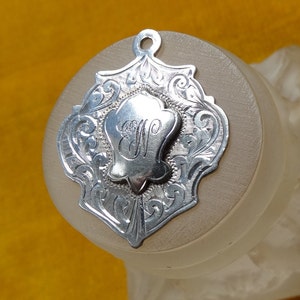 May include: Silver pendant featuring an intricate design and a monogrammed initial "V" at its centre.
