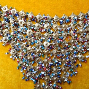 Puede incluir: Un collar de oro con un diseño en cascada de pequeñas cuentas de colores y abalorios de metal. El collar está sobre un fondo amarillo.