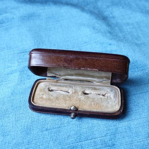 May include: A brown leather ring box with a white velvet interior and two compartments. The box is open and the compartments are empty.