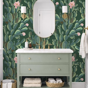 May include: Wallpaper featuring a repeating pattern of green prickly pear cactus pads and light pink flowers on a dark forest green background. The design covers the wall behind a sage green vanity cabinet with gold hardware and a white countertop.