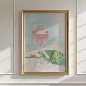 K&ouml;nnte beinhalten: Gerahmtes Gem&auml;lde mit einem Weinglas mit rosa Wein, einem gr&uuml;nen Pickleball-Schl&auml;ger und einem gr&uuml;nen Pickleball. Das Kunstwerk hat einen goldenen Rahmen und einen hellblauen und wei&szlig;en Hintergrund.