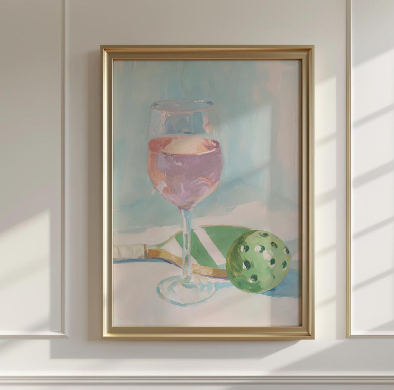 K&ouml;nnte beinhalten: Gerahmtes Gem&auml;lde mit einem Weinglas mit rosa Wein, einem gr&uuml;nen Pickleball-Schl&auml;ger und einem gr&uuml;nen Pickleball. Das Kunstwerk hat einen goldenen Rahmen und einen hellblauen und wei&szlig;en Hintergrund.