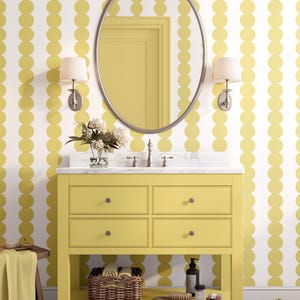 Puede incluir: Un mueble de baño amarillo con cuatro cajones, una encimera de mármol y un espejo ovalado plateado. La pared está decorada con un patrón de lunares blancos y amarillos. Una cesta tejida y accesorios están en el estante de abajo.
