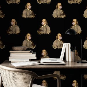 May include: Black wallpaper with a repeating pattern of Benjamin Franklin portraits blowing bubble gum. Each portrait is framed by a gold laurel wreath. The image includes a desk with books, a lamp, and a chair.