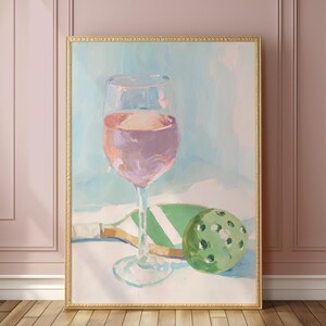 K&ouml;nnte beinhalten: Ein &Ouml;lgem&auml;lde zeigt ein Weinglas mit rosa Fl&uuml;ssigkeit, einen gr&uuml;nen Pickleball und einen Schl&auml;ger. Das Kunstwerk ist goldgerahmt und vor einem hellblauen und wei&szlig;en Hintergrund platziert. Der Stil ist impressionistisch.