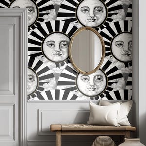 May include: A wall decorated with wallpaper featuring a repeating pattern of sun faces with black and white rays. An oval gold-framed mirror hangs on the wall. A wooden bench with white pillows sits below the mirror.
