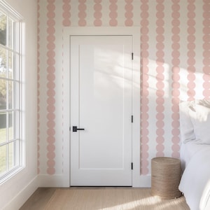 May include: A bedroom interior featuring a white door with a black handle, a window, and walls adorned with a pink polka dot pattern. A woven stool sits near a bed with white linens, creating a bright and airy space.