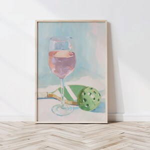 K&ouml;nnte beinhalten: Gerahmtes Gem&auml;lde, das ein Weinglas mit rosa Fl&uuml;ssigkeit, einen gr&uuml;nen Pickleball und einen Pickleball-Schl&auml;ger zeigt. Das Kunstwerk hat eine weiche Pastellfarbpalette mit einem hellblauen und wei&szlig;en Hintergrund.