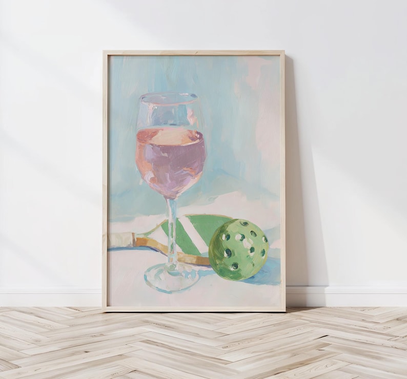 K&ouml;nnte beinhalten: Gerahmtes Gem&auml;lde, das ein Weinglas mit rosa Fl&uuml;ssigkeit, einen gr&uuml;nen Pickleball und einen Pickleball-Schl&auml;ger zeigt. Das Kunstwerk hat eine weiche Pastellfarbpalette mit einem hellblauen und wei&szlig;en Hintergrund.