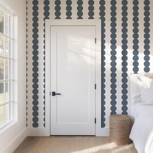 Può includere: Un interno bianco con una porta bianca chiusa, una finestra e un letto. Le pareti sono decorate con un motivo ripetuto di cerchi blu scuro. Un piccolo sgabello cilindrico intrecciato è posizionato vicino al letto.