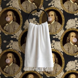 May include: A white, fringed hand towel hangs from a black metal ring. The background is a repeating pattern of portraits of Benjamin Franklin blowing bubble gum, set against a black floral wallpaper.