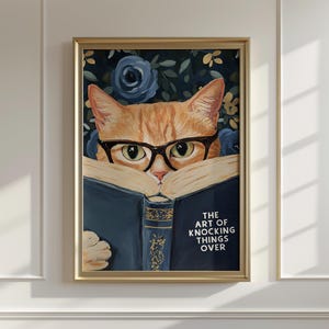 Könnte beinhalten: Gerahmter Kunstdruck mit einer orangefarbenen Katze mit Brille, die ein Buch liest. Auf dem offenen Buch steht "THE ART OF KNOCKING THINGS OVER". Der Hintergrund zeigt ein Blumenmuster mit blauen Rosen.