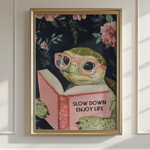May include: Framed art print featuring a turtle wearing pink glasses and reading a book. The book is open, displaying the text "SLOW DOWN ENJOY LIFE". The background has a dark floral pattern with pink flowers.