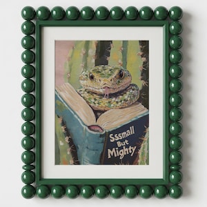Op de afbeelding: Een ingelijste kunstdruk met een groene slang die op een open boek voor een cactus rust. De boekomslag bevat de tekst Sssmall But Mighty. De lijst is donkergroen met een decoratieve, afgeronde, kraalachtige textuur langs de randen.