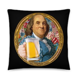 Puede incluir: Una almohada negra con un retrato enmarcado en oro de un hombre sosteniendo un vaso de cerveza. El retrato está sobre un fondo de flores coloridas.