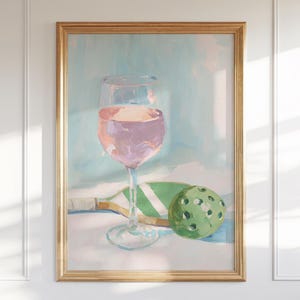 K&ouml;nnte beinhalten: Gerahmtes &Ouml;lgem&auml;lde, das ein Weinglas mit rosa Fl&uuml;ssigkeit, einen gr&uuml;nen Pickleball und einen Pickleball-Schl&auml;ger zeigt. Der Hintergrund ist ein weiches Blau und Wei&szlig;. Das Kunstwerk hat eine leichte, luftige Ausstrahlung.
