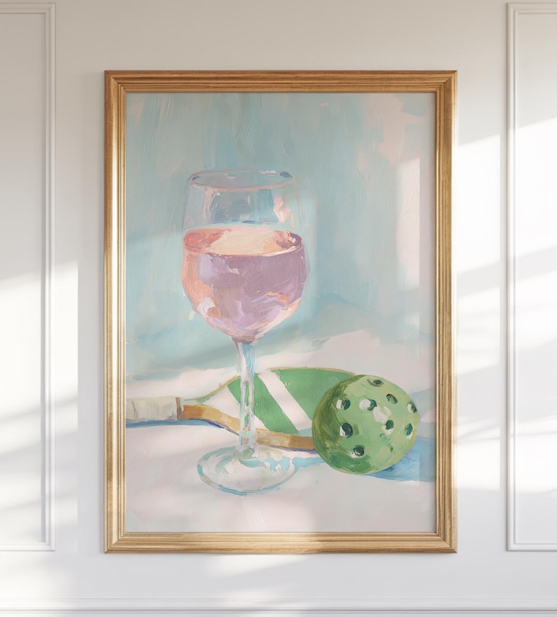 K&ouml;nnte beinhalten: Gerahmtes &Ouml;lgem&auml;lde, das ein Weinglas mit rosa Fl&uuml;ssigkeit, einen gr&uuml;nen Pickleball und einen Pickleball-Schl&auml;ger zeigt. Der Hintergrund ist ein weiches Blau und Wei&szlig;. Das Kunstwerk hat eine leichte, luftige Ausstrahlung.