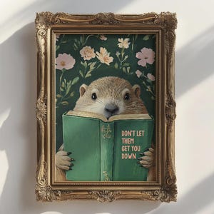 May include: An artwork in a gold frame depicts a groundhog reading a green book. The book's text reads "DON'T LET THEM GET YOU DOWN." The background is dark green with floral patterns. The frame has an ornate design.