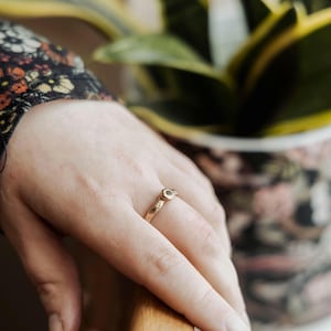 Op de afbeelding: Een gouden ring met een kleine, ronde, groene edelsteen in het midden. De ring wordt aan een vinger gedragen.